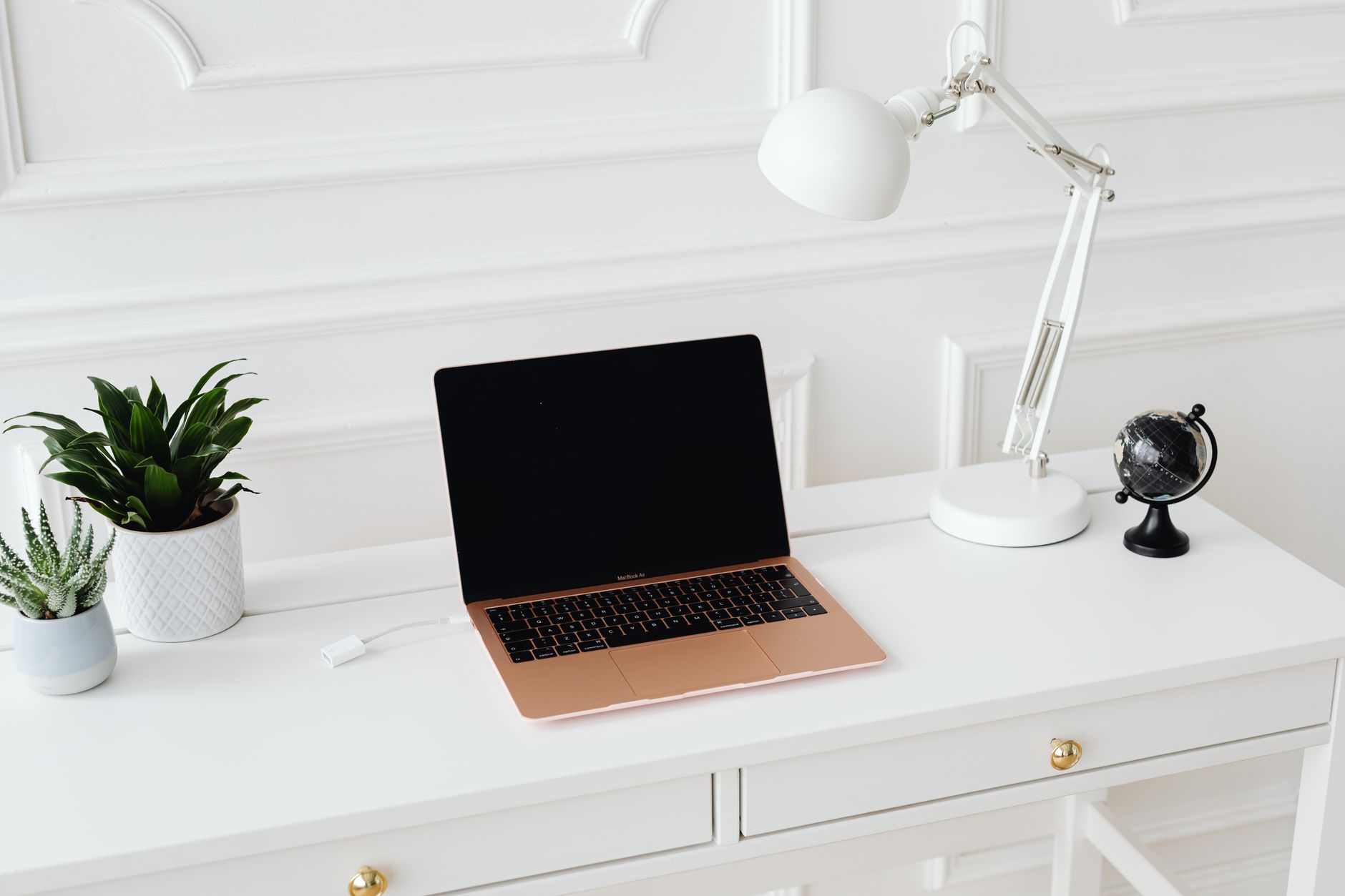 Bureau blanc lumineux avec un ordinateur portable ouvert, une lampe articulée, un petit globe et des plantes en pot