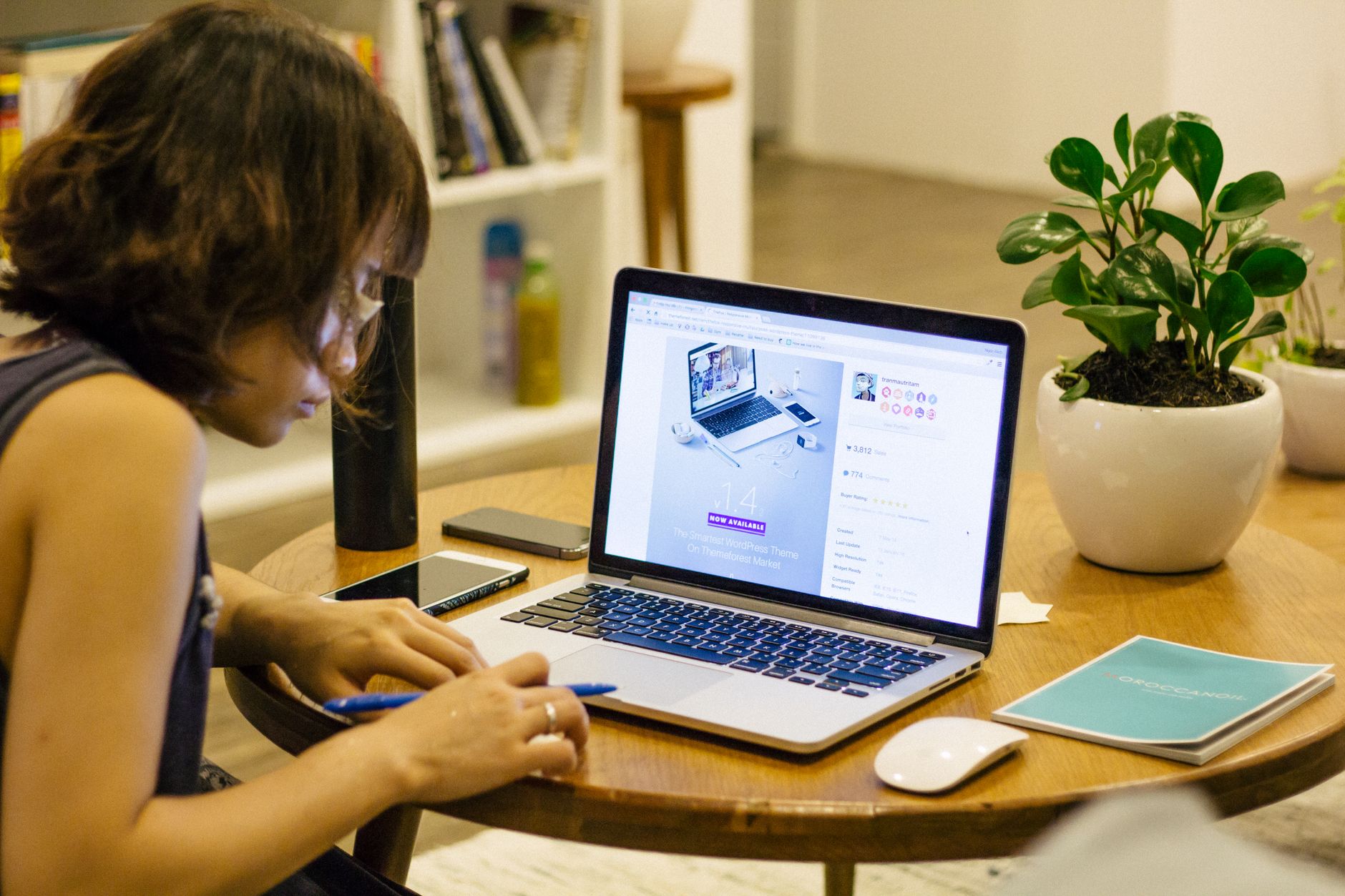 Femme de profil concentrée devant un ordinateur portable, un smartphone posé à côté, sur une table ronde près d’une plante