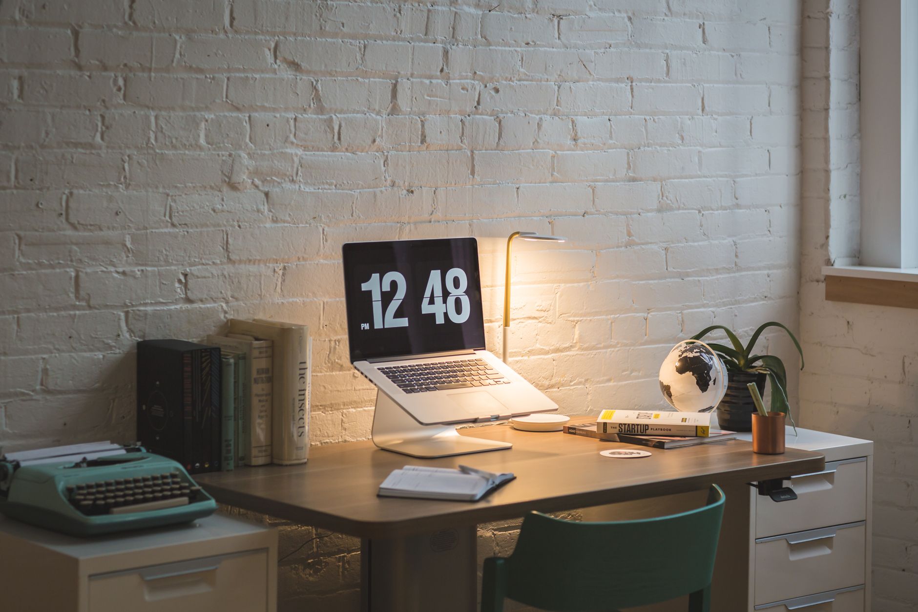 Bureau éclairé par une lampe devant un mur de briques blanches, avec un ordinateur portable sur support, des livres et un carnet